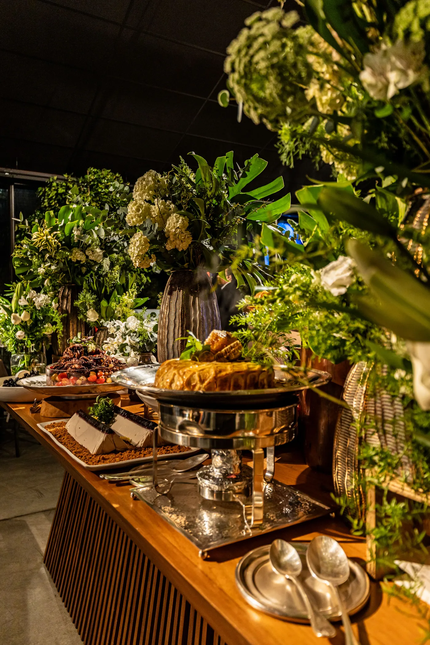 Mesa de buffet em madeira com réchaud de prata e arranjos de folhagens em evento noturno